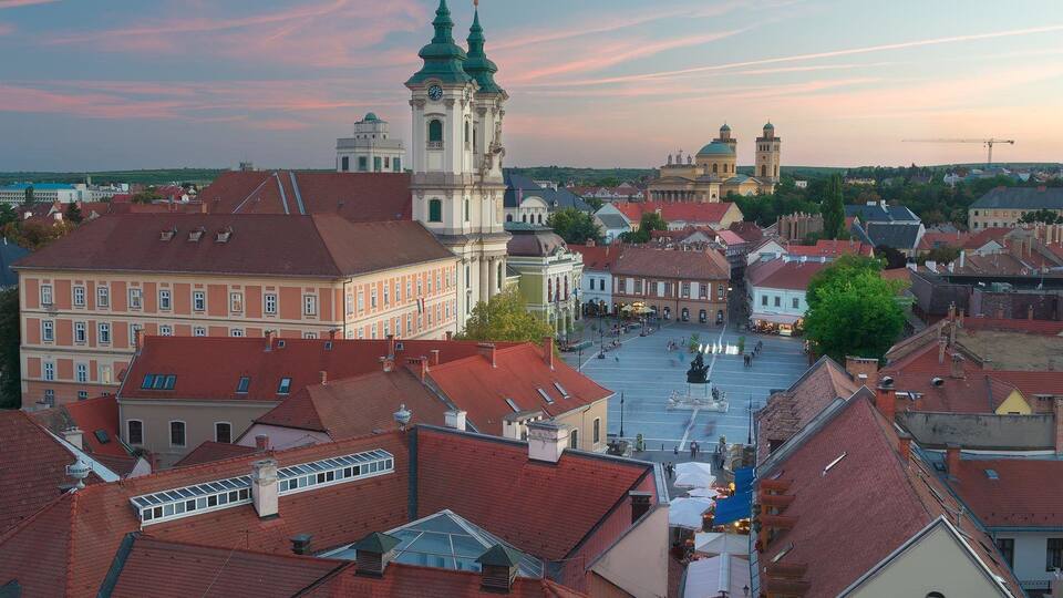 A cityscape with a prominent church with green spires, a central square with fountains, and surrounding buildings with red roofs.