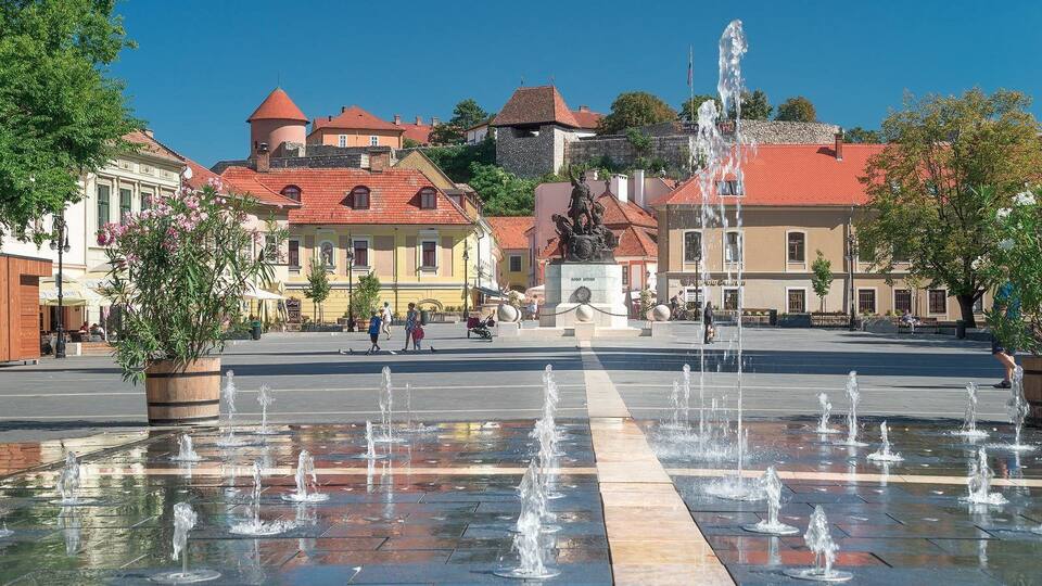 A public square with a fountain, historic buildings, and a statue.