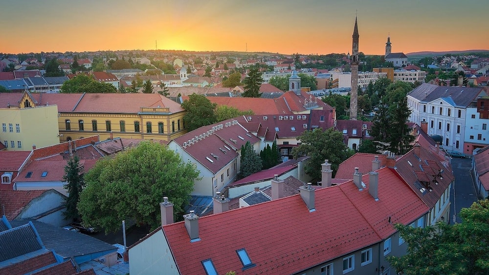 Bybillede med røde tage, et markant kirketårn og en solnedgang i baggrunden.