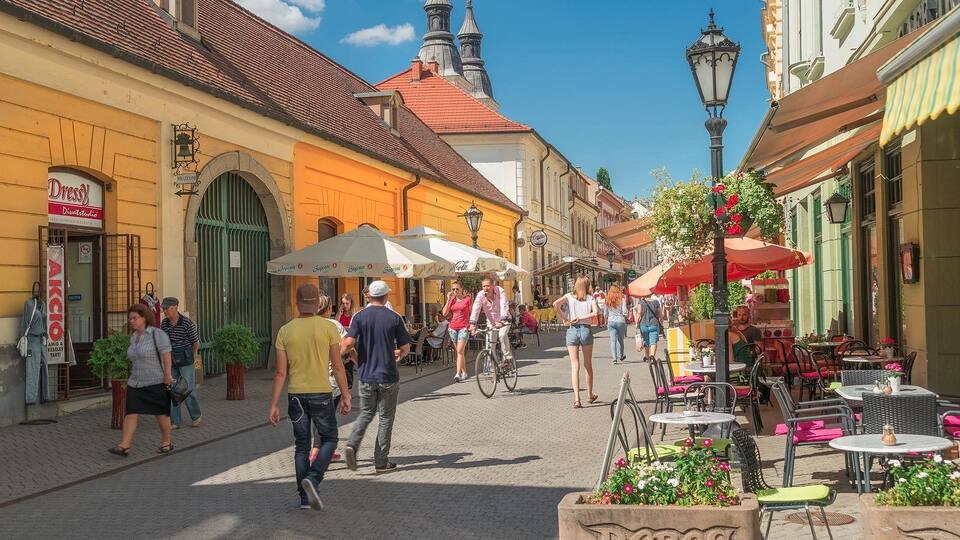 A cobblestone street lined with cafes and shops, a church with a red roof, and pedestrians walking and cycling.