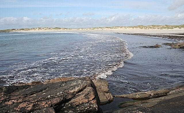 A rocky shoreline with waves crashing onto the beach and a distant sandy dune.