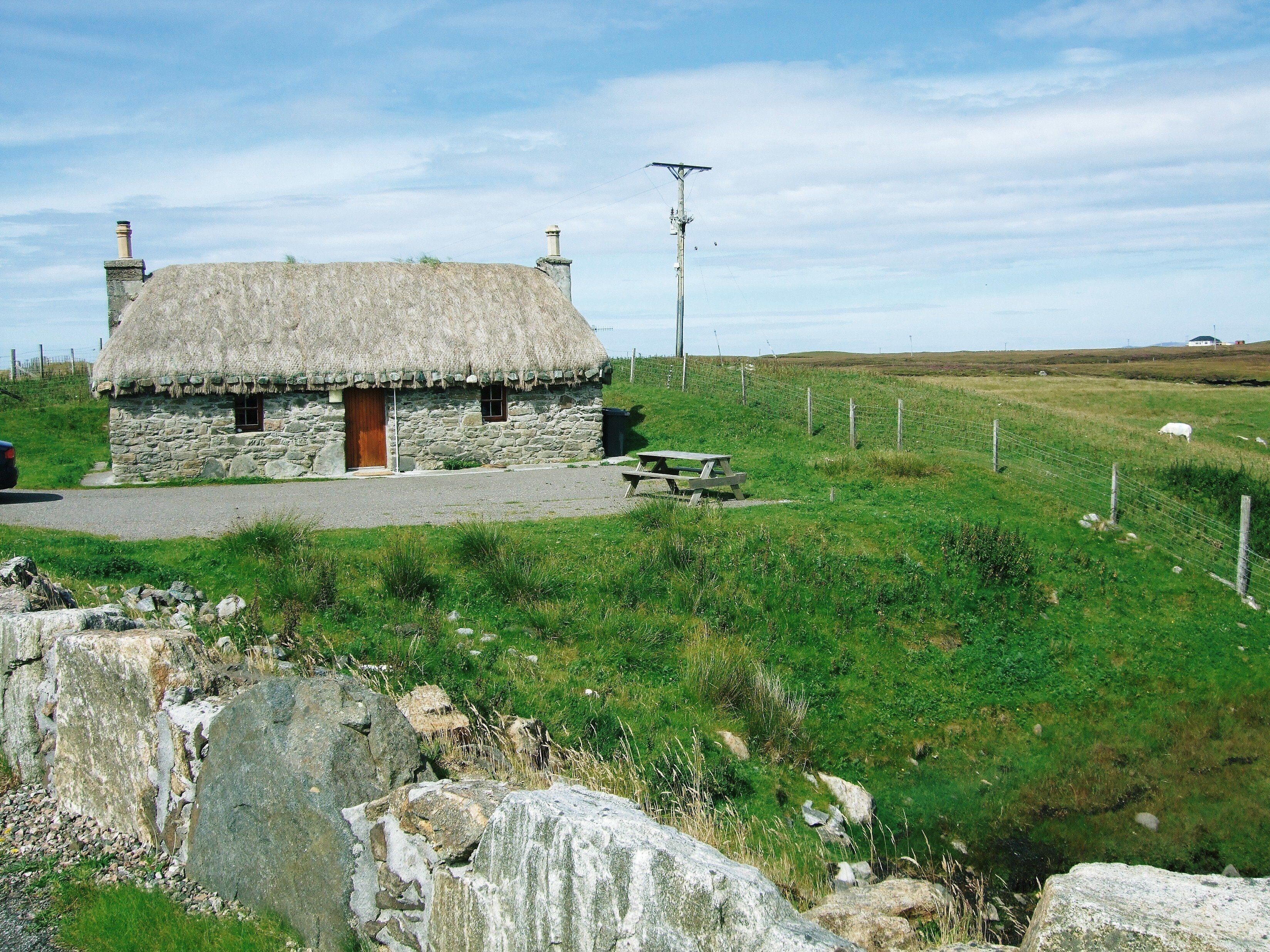 A stone house with a thatched roof, a picnic table, and a grassy field with a fence.