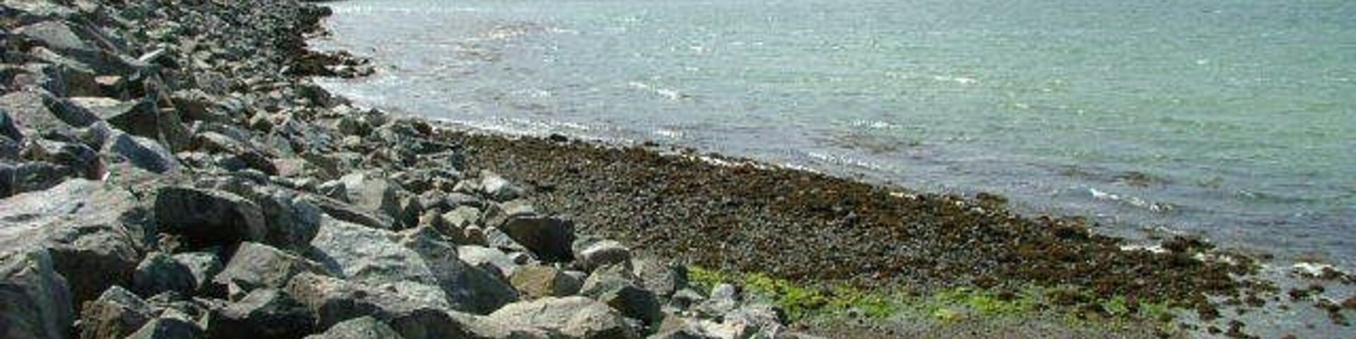 Una costa rocosa con un muelle que se adentra en aguas tranquilas, una cadena montañosa distante y un cielo nublado.