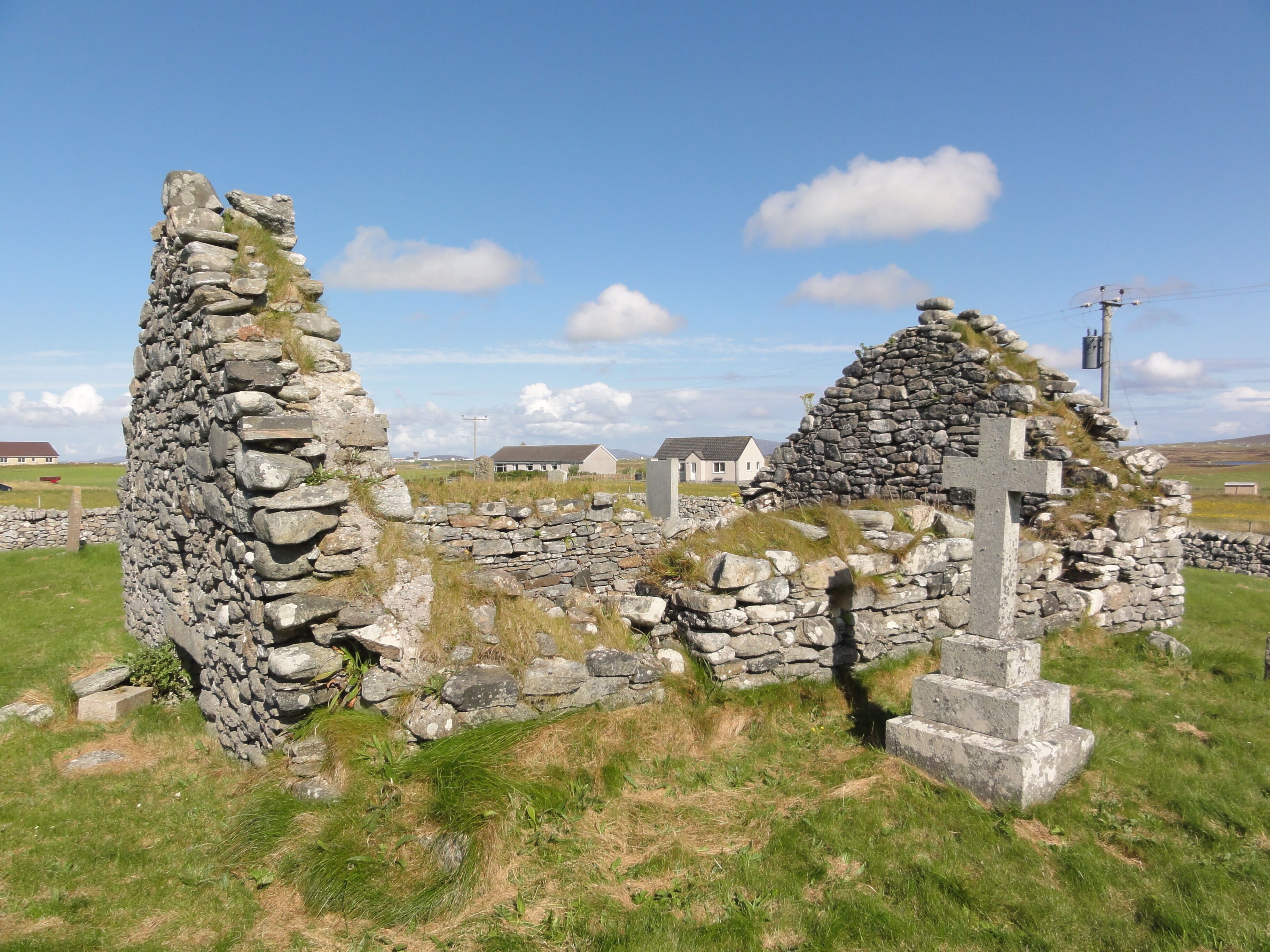 A stone cross and ruins in a grassy field with houses in the background.