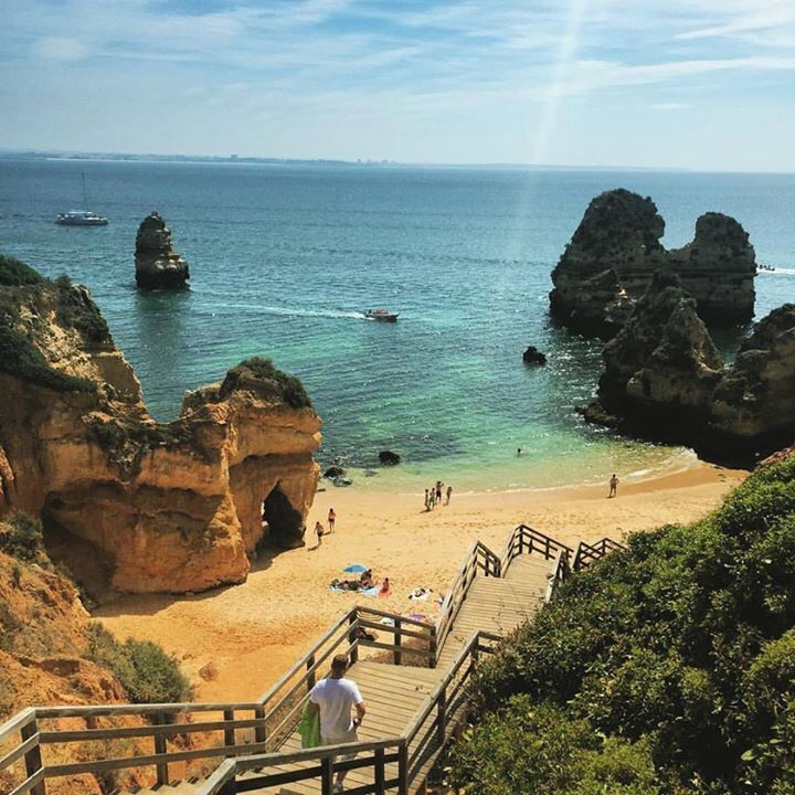 A coastal beach with rocky formations, a wooden staircase, and clear blue water.