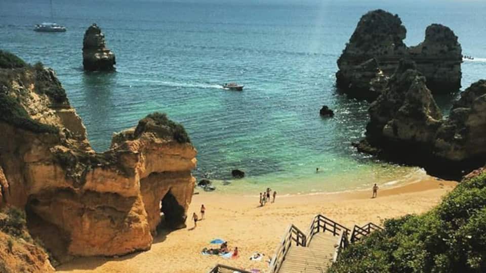 A coastal beach with rocky formations, a wooden staircase, and clear blue water.
