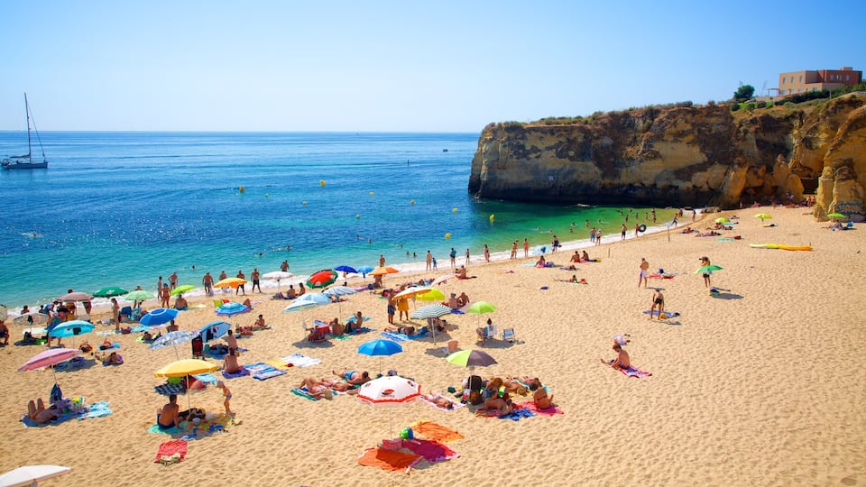 A crowded beach with people sunbathing and swimming, colorful umbrellas, and a rocky cliff in the background.