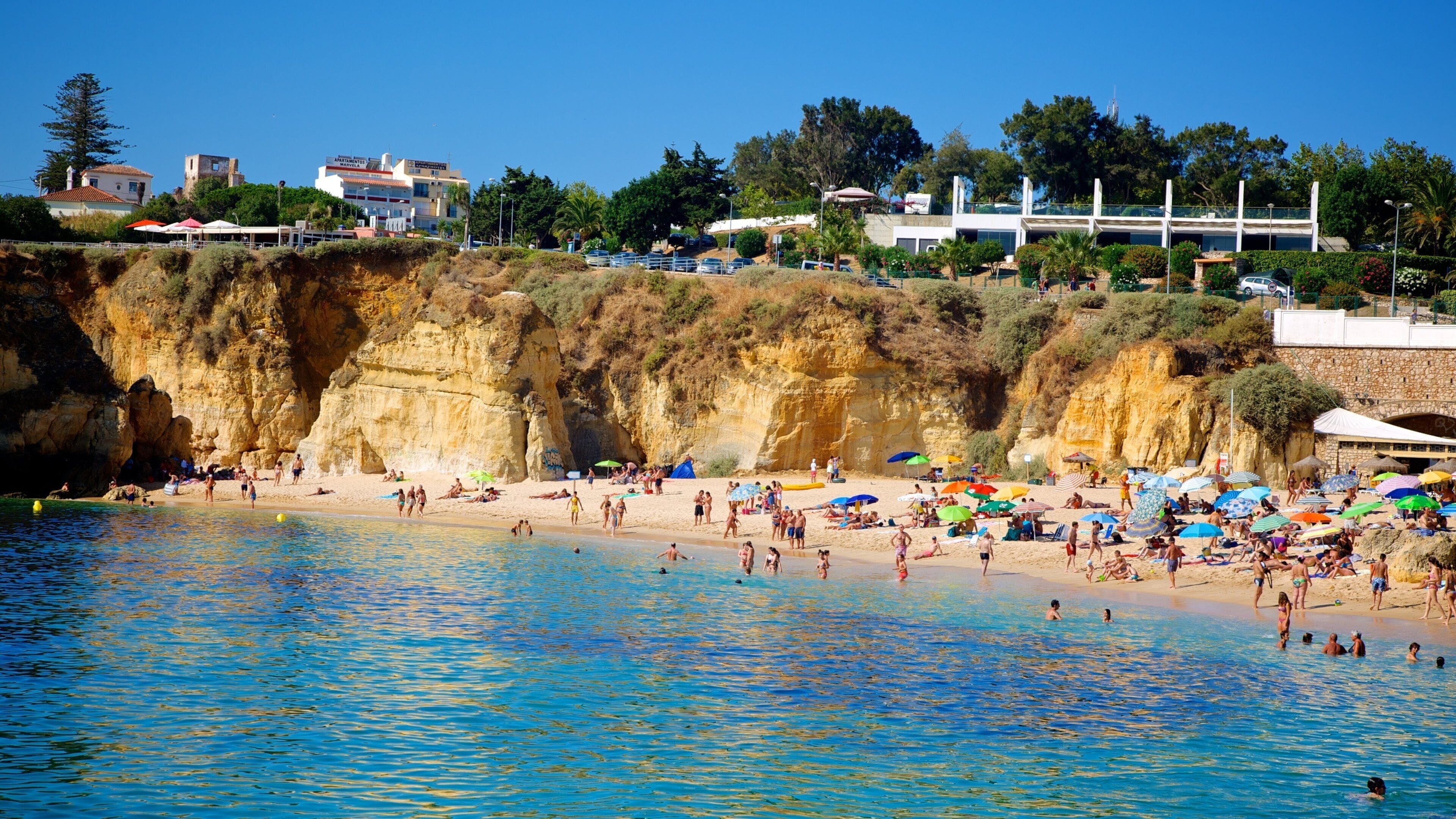 A crowded beach with clear blue water, rocky cliffs, and buildings in the background.