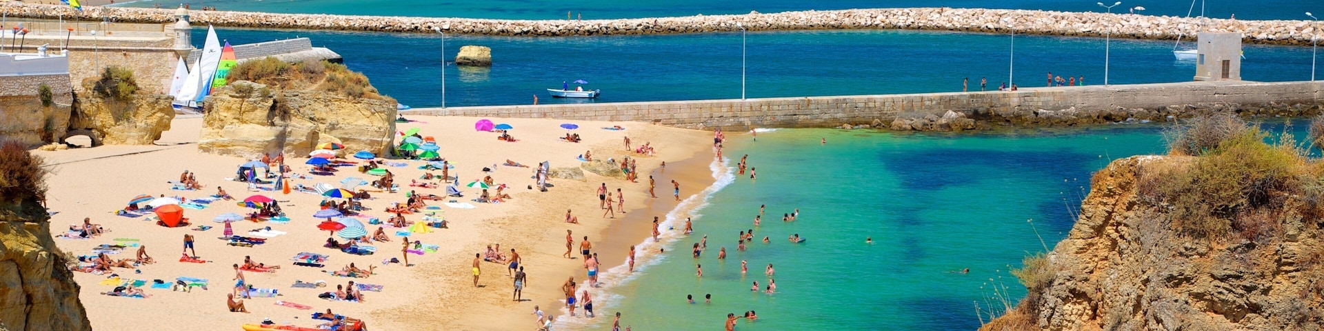 A crowded beach with clear turquoise water, rocky cliffs, and a distant mountain range.