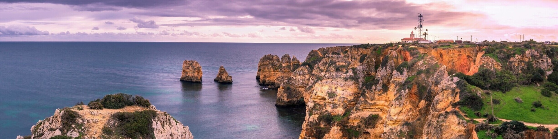 Paisagem costeira com falésias rochosas, um farol e mar azul cristalino.