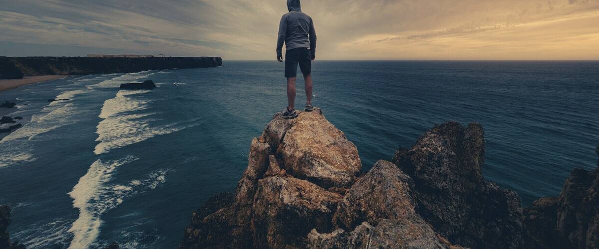 Une personne se tient sur un promontoire rocheux, surplombant une plage et l’océan, sous un ciel nuageux.