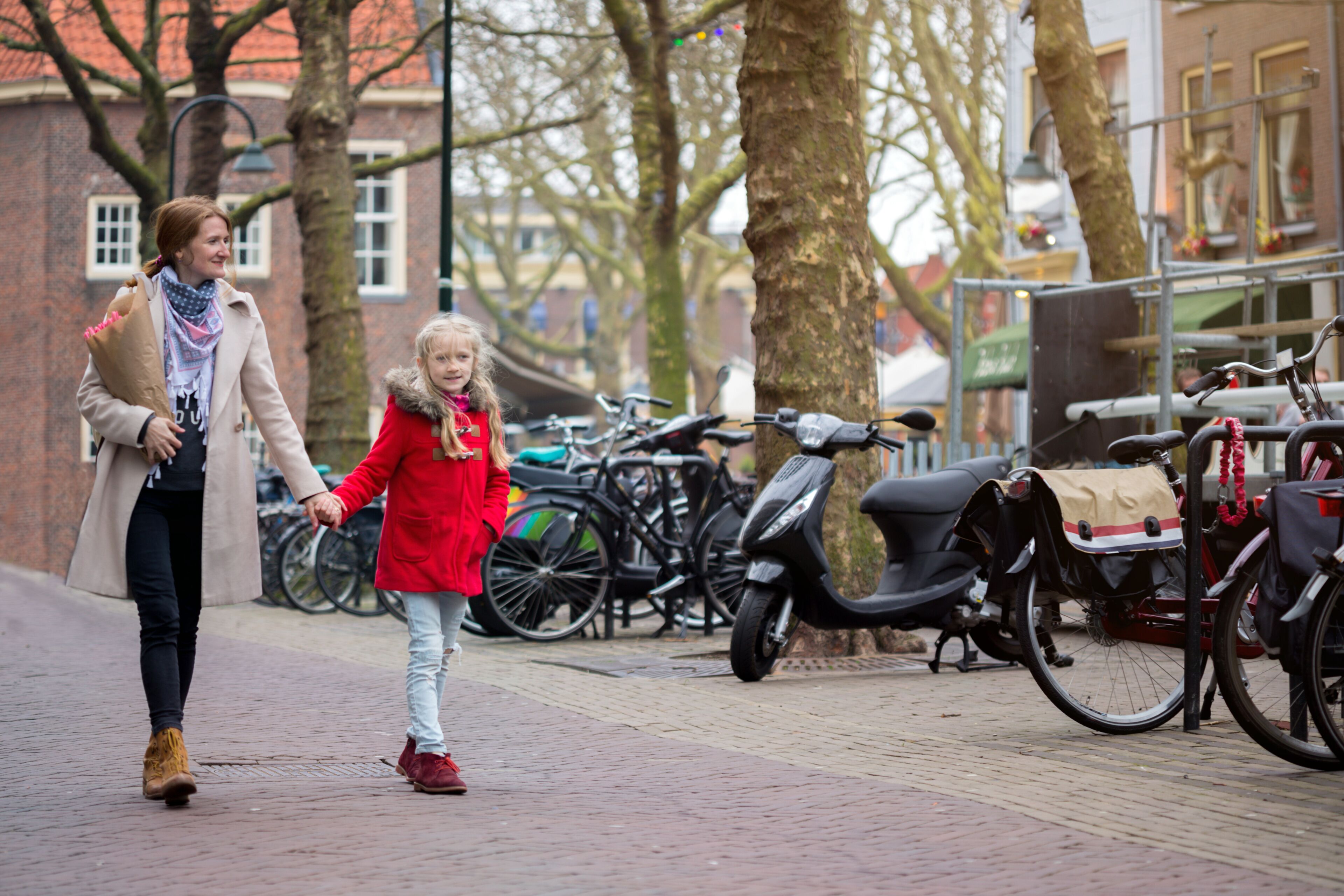 Uma mulher e uma criança caminham em um caminho de tijolos, de mãos dadas, com bicicletas e scooters estacionadas ao lado.