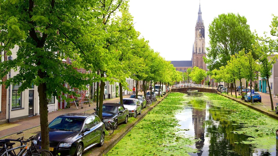 A canal lined with trees and parked cars, a bridge, and a church tower in the background.