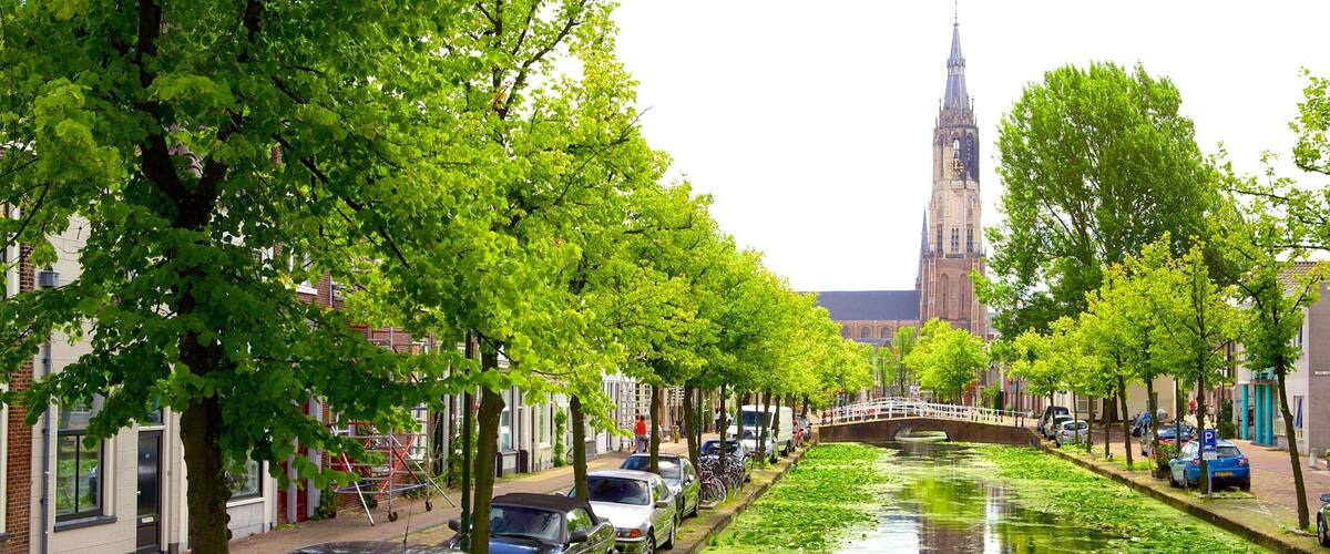 A canal lined with trees and parked cars, a bridge, and a church tower in the background.