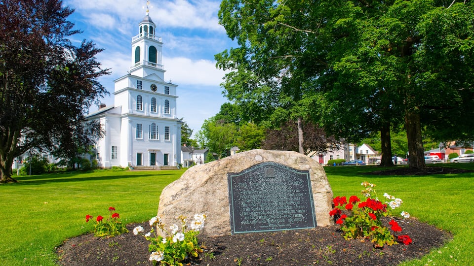 A white church with a steeple, surrounded by a grassy area with a stone monument and flowering plants.