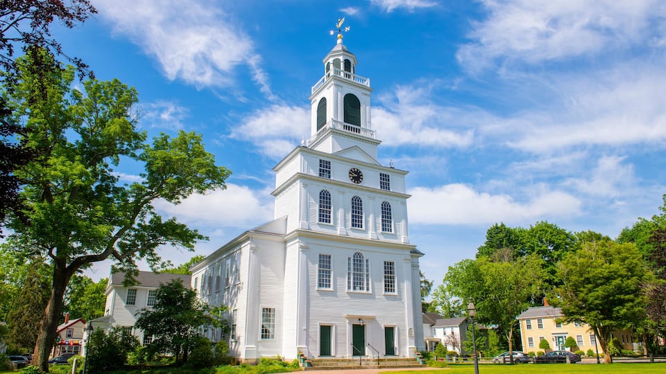 A white church with a bell tower and a clock, surrounded by green grass and trees.