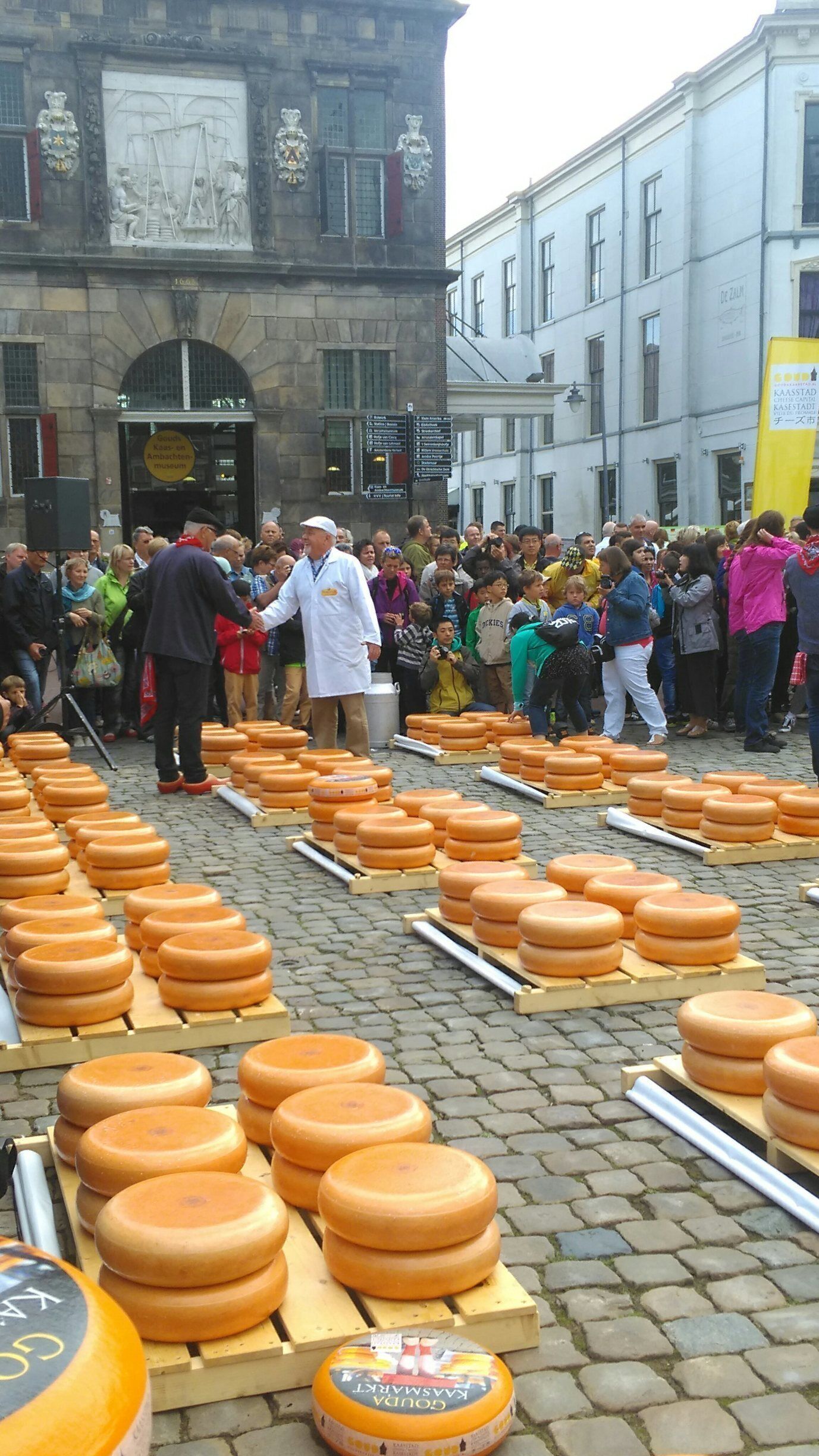 A crowd of people gathered around a display of cheese wheels in an outdoor market setting.