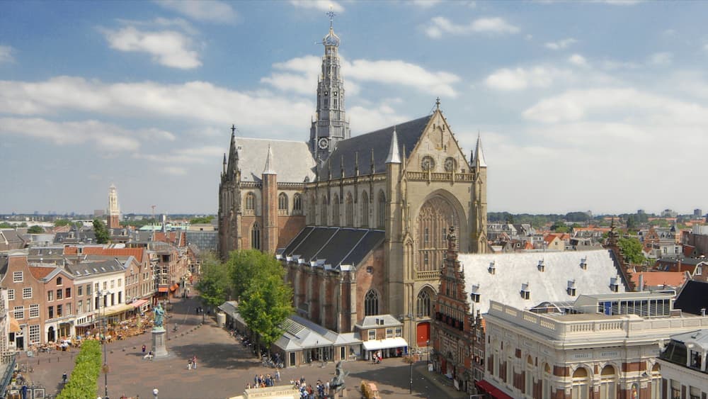 Une église historique, dotée d’un flèche imposante et d’une tour horloge, qui s’élève au milieu d’une place animée, bordée de cafés et fréquentée par de nombreux piétons.