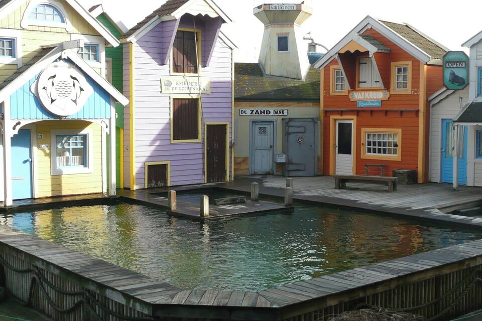 Un colorido paseo marítimo con edificios que muestran las indicaciones 'SMEDERJ DE IJZEREN' y 'VAT KRUID'.