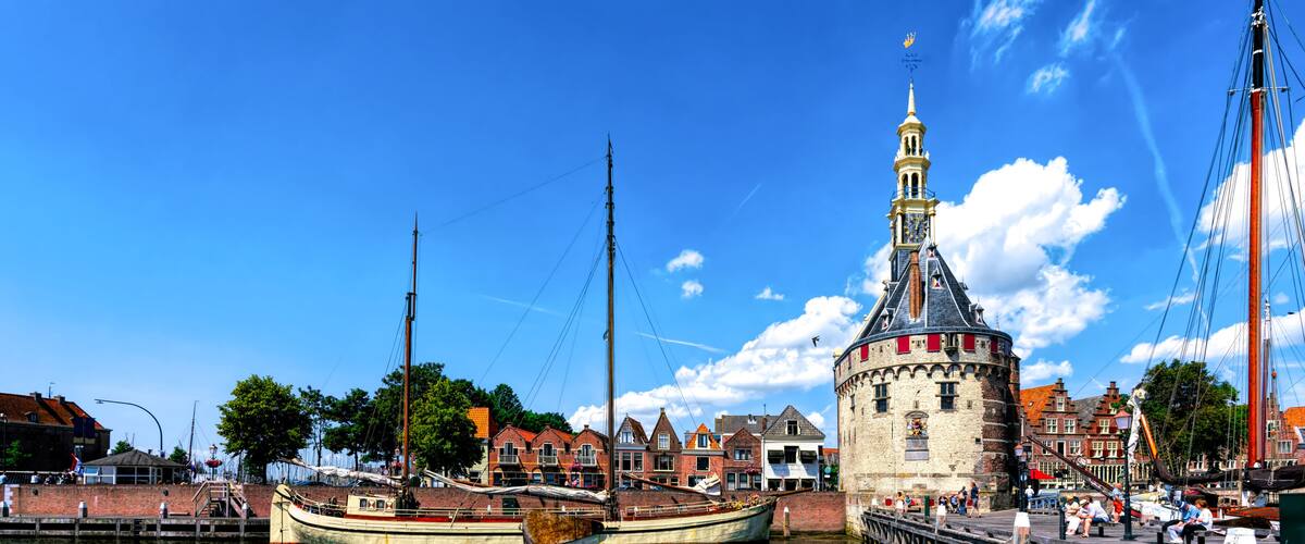 A historic tower with a clock, a traditional boat docked at a pier, and a waterfront with buildings.