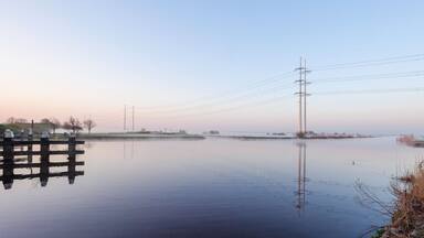 Un río tranquilo con un muelle de madera, líneas eléctricas y árboles distantes.