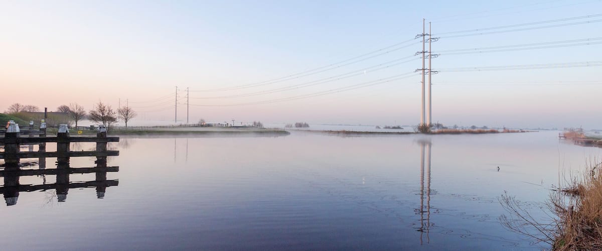 A calm river with a wooden dock, power lines, and distant trees.
