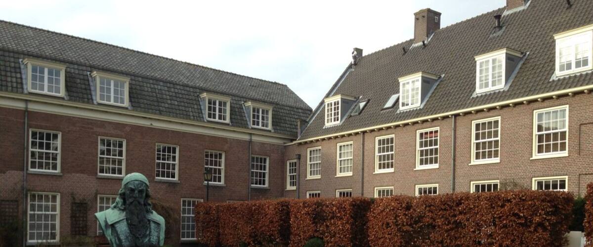 A statue of Jan Amos Comenius in a garden with a brick building in the background.