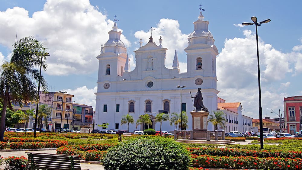 A white church with three towers, surrounded by a well-maintained garden with red flowers and palm trees.