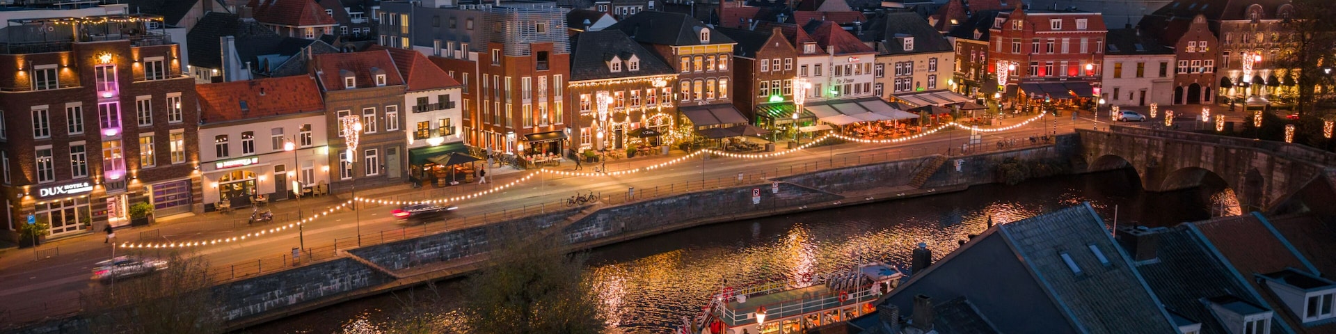 Une ville riveraine au crépuscule, avec des bâtiments illuminés, un clocher d’église imposant et un pont.