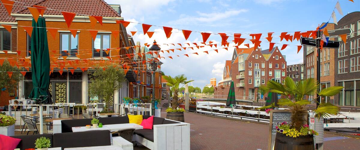 A riverside café with outdoor seating, orange triangular flags, and traditional European buildings in the background.