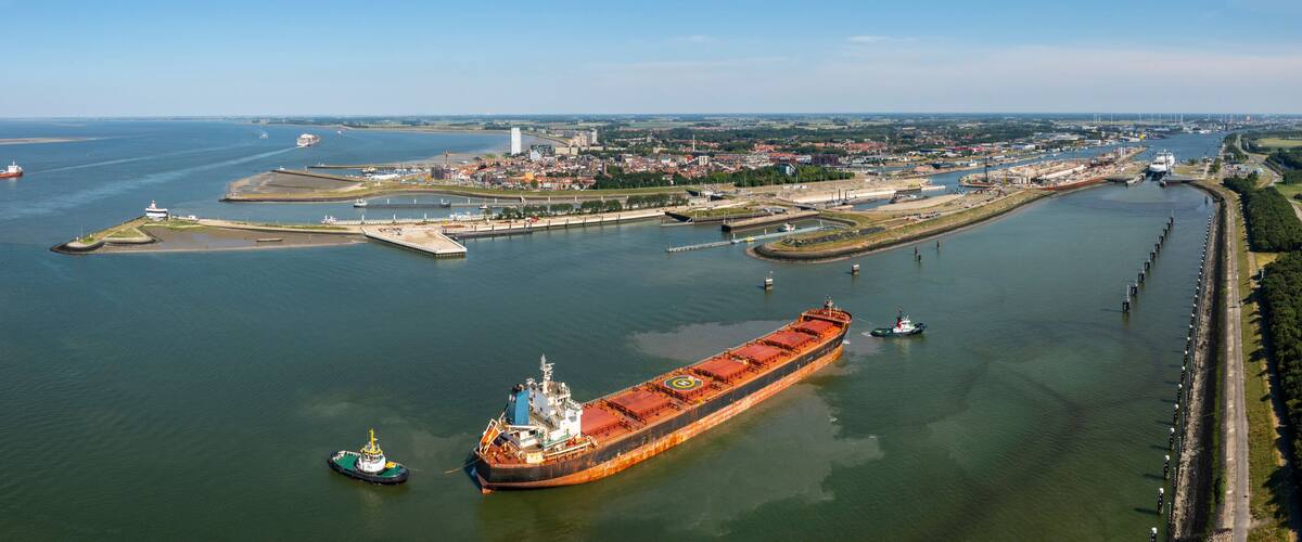 A large cargo ship docked at a port with a tugboat nearby, surrounded by a cityscape and a river.