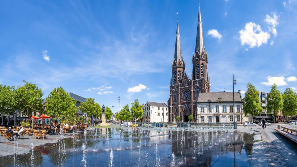 A historic church with twin spires, a fountain in the foreground, and a café-lined square.