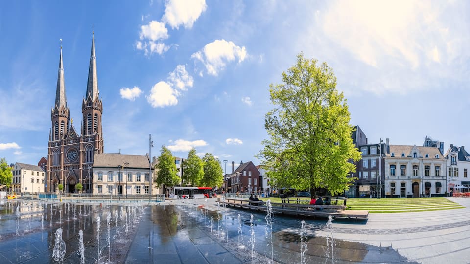 Eine historische Kirche mit zwei Türmen, einem Brunnen und einem gepflegten Platz mit Bäumen und Gebäuden.