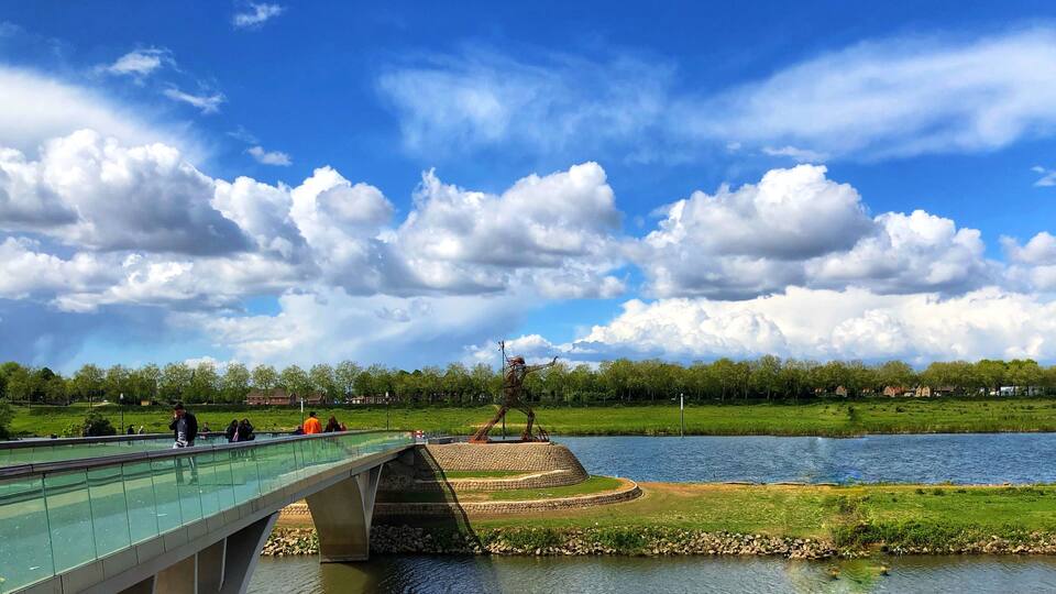 A bridge over a river with a statue in the center, surrounded by greenery and houses in the background.