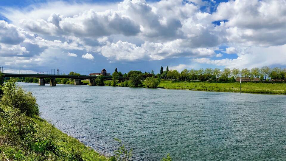 A river with a bridge, greenery, and a partly cloudy sky.