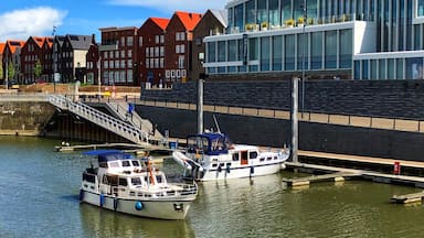 Un canal où sont amarrés deux bateaux à un quai, un bâtiment moderne doté de grandes fenêtres, et, au fond, des maisons traditionnelles aux toits rouges.