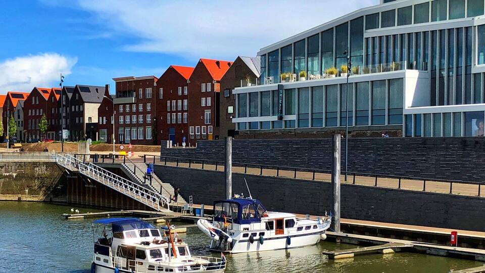 A canal with two boats docked at a pier, a modern building with large windows, and traditional red-roofed houses in the background.