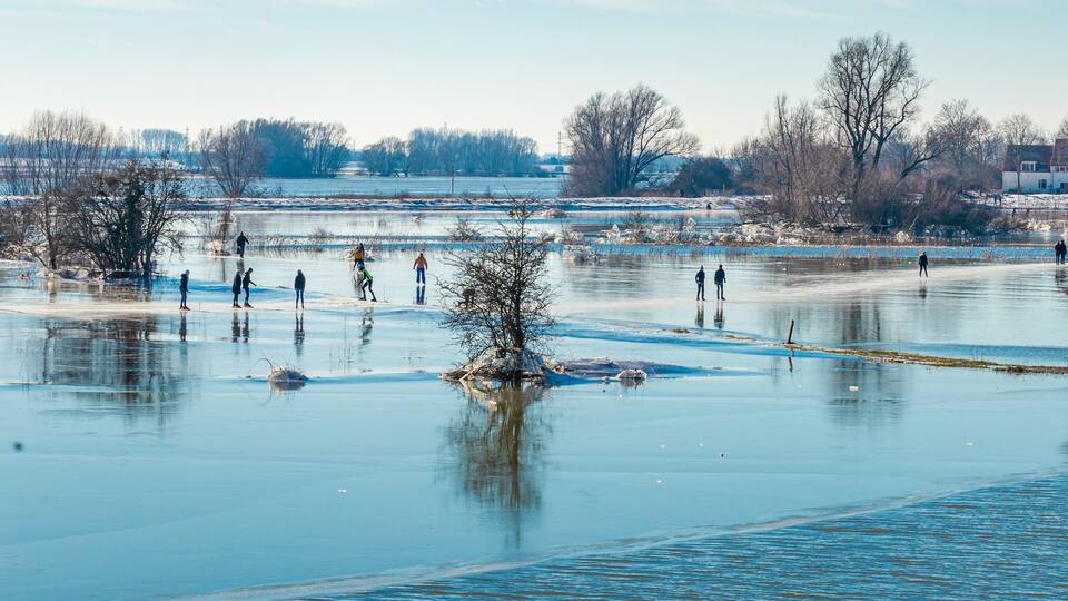 A frozen river with people ice skating, surrounded by leafless trees and industrial buildings.