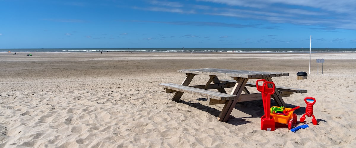 A sandy beach with a wooden picnic table and red beach toys.