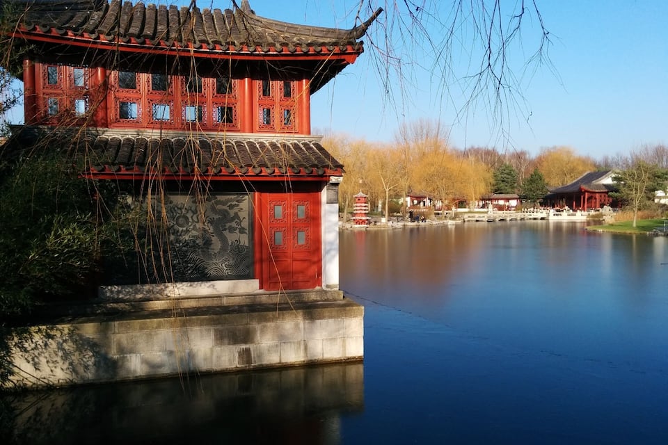 Um edifício tradicional chinês com portas vermelhas e desenhos complexos nas janelas, localizado à beira de um lago tranquilo com um pagode ao fundo.
