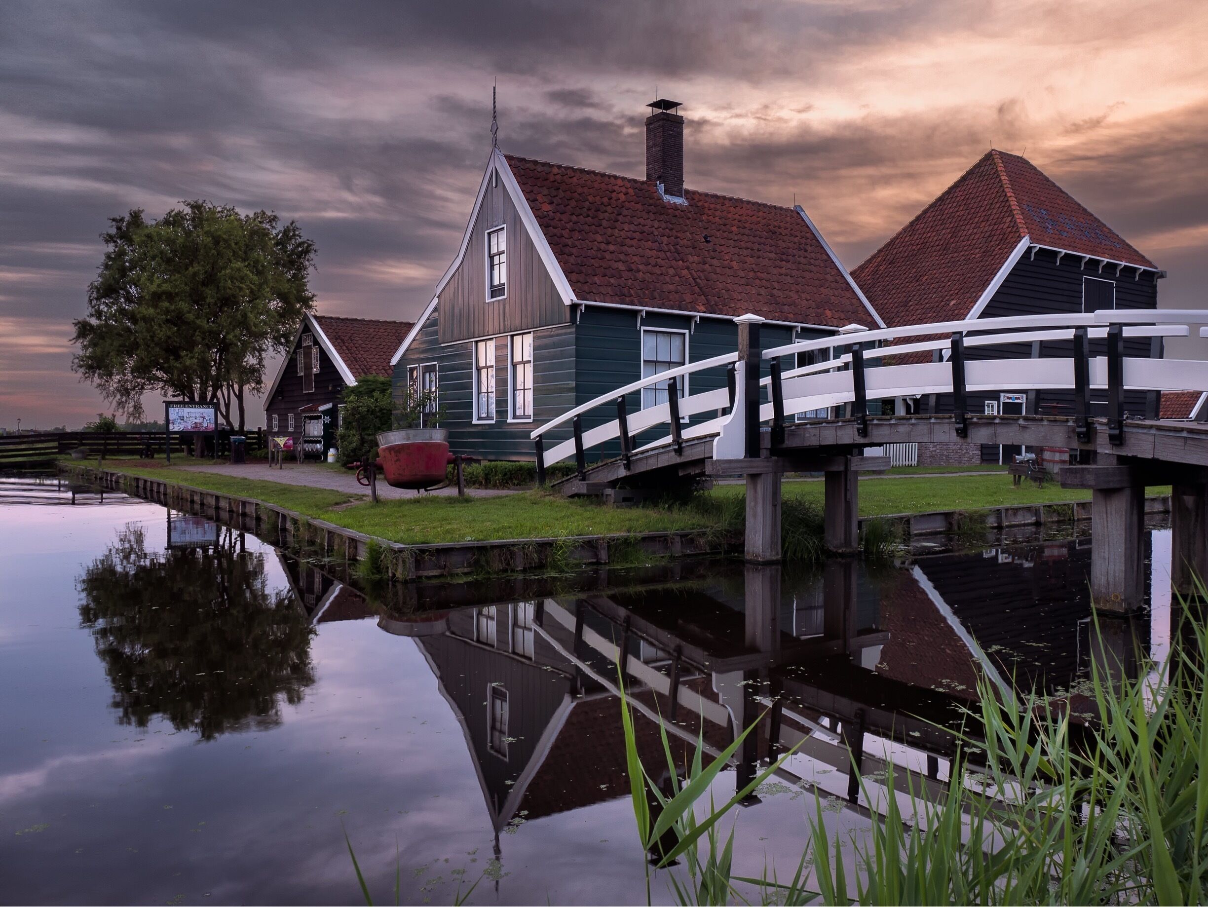 A canal with a wooden bridge, houses with red roofs, and a calm water surface reflecting the sky.