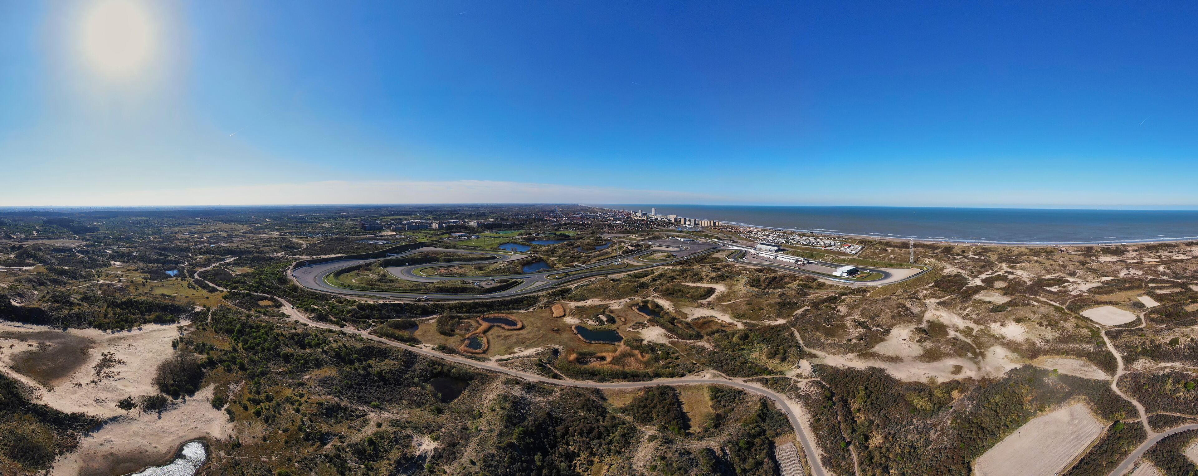 An aerial view of a coastal area with a large racetrack, a marina, and a cityscape in the background.