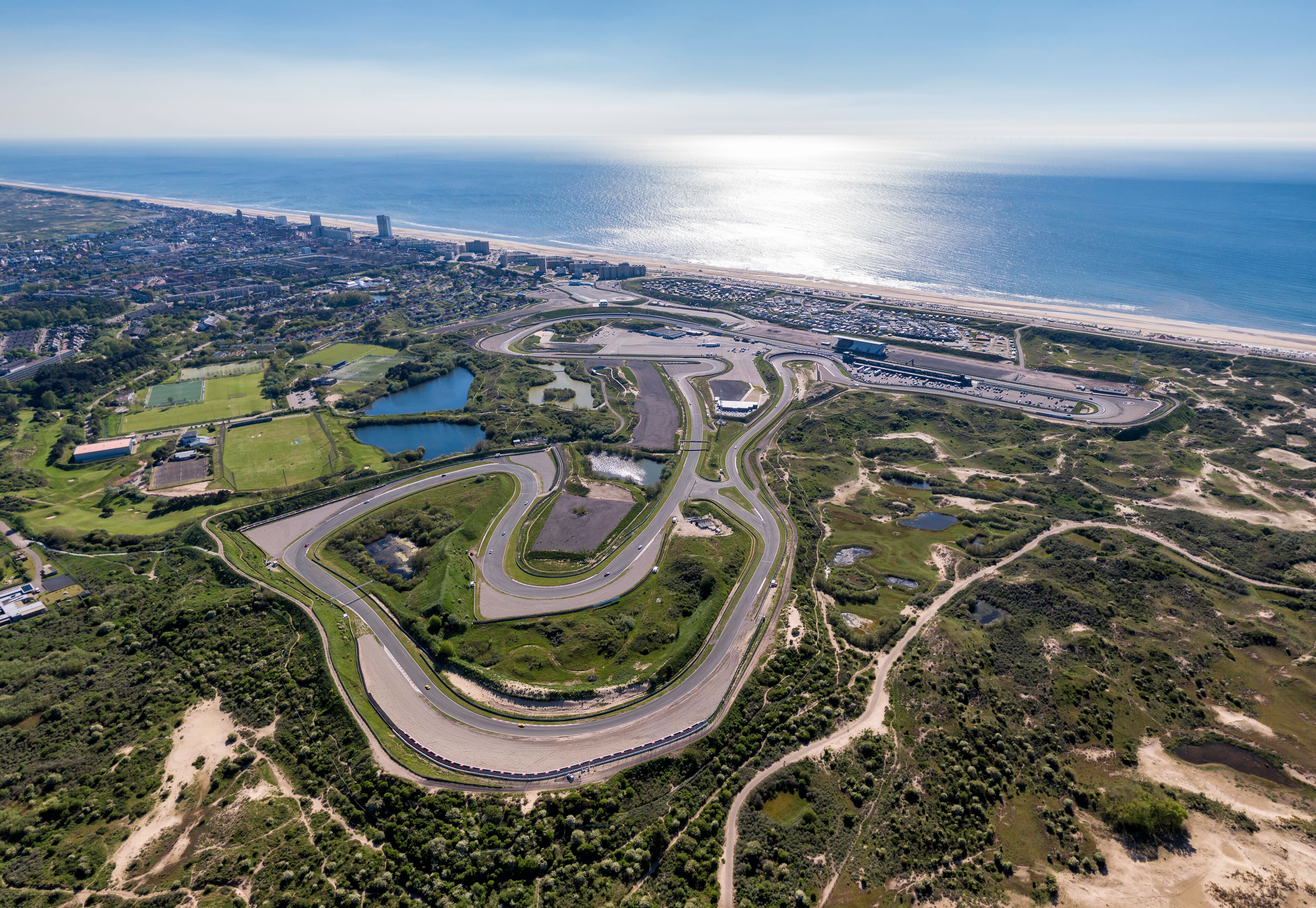 An aerial view of a coastal racing circuit with multiple turns, a beach, and a cityscape in the background.