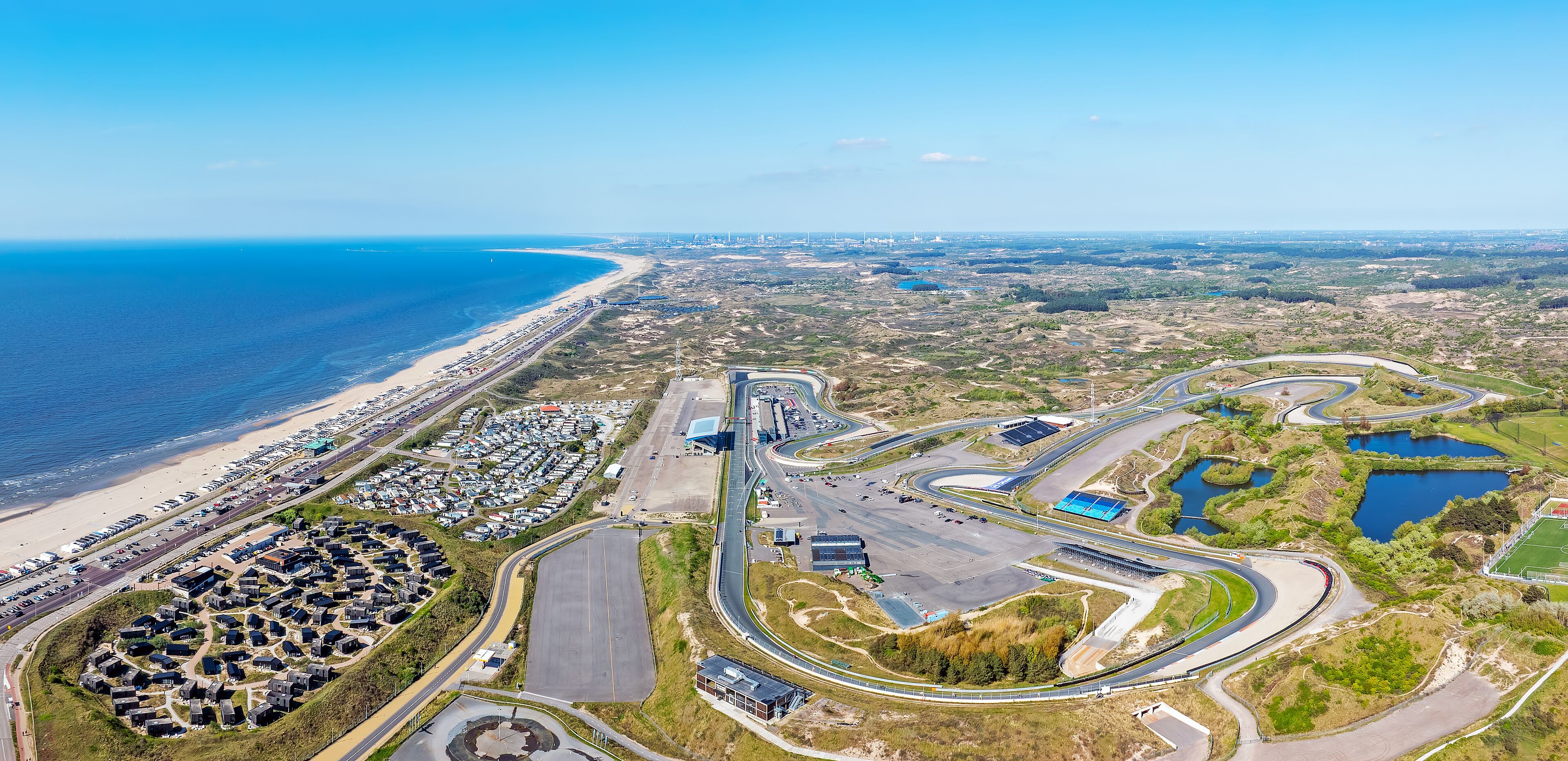 An aerial view of a coastal area with a beach, parking lots, and a racetrack.