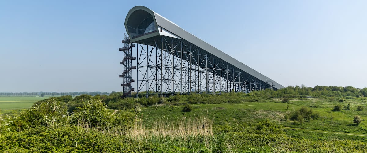 A large, modern, arched building with a metal framework, set in a lush green landscape with rolling hills in the background.