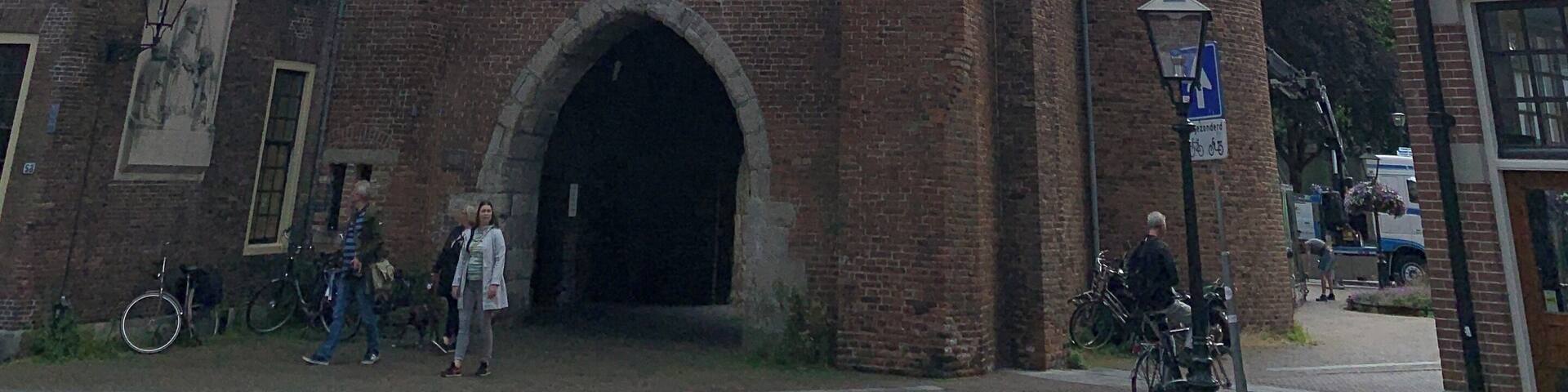A historic brick tower with a clock and pointed roof, surrounded by traditional buildings.