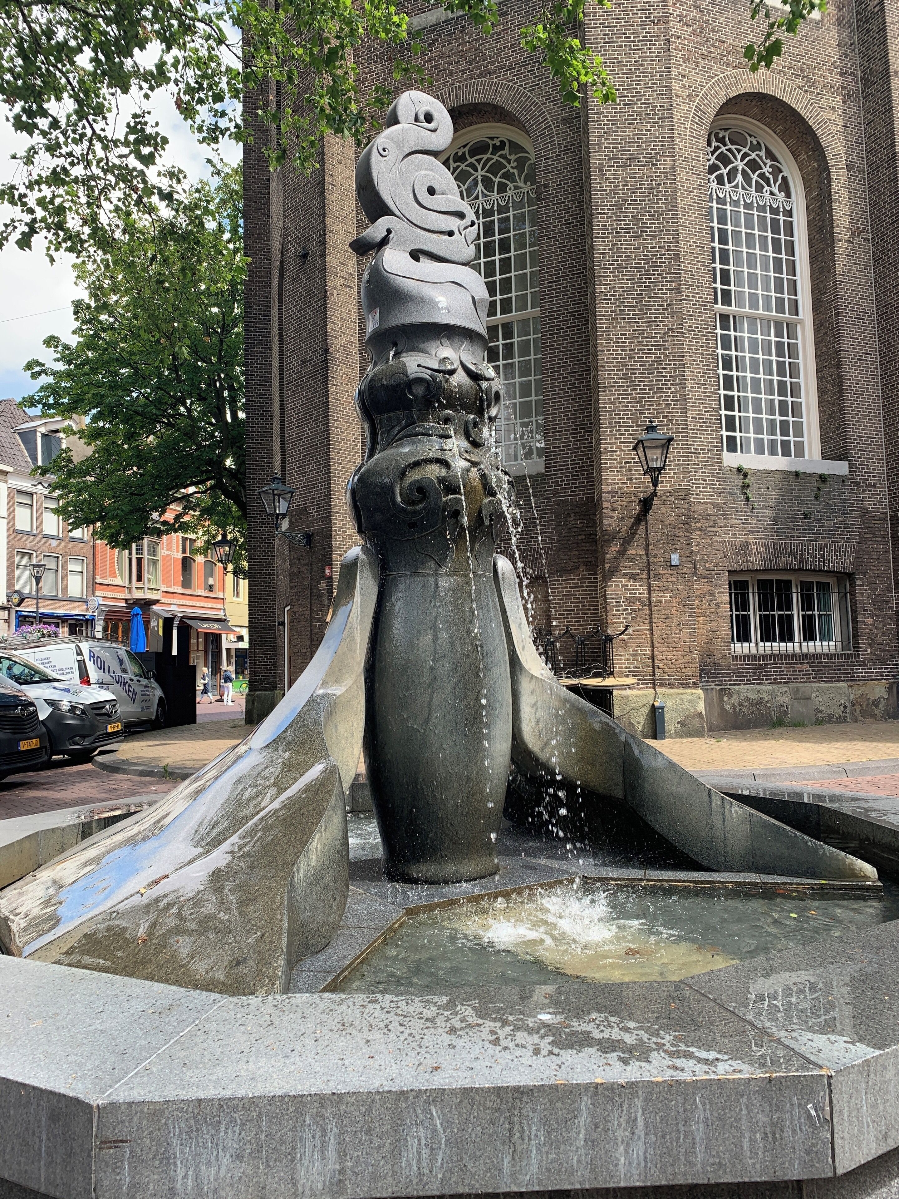 Una fuente de piedra con agua brotando de su centro, frente a un edificio de ladrillo con ventanales arqueados.