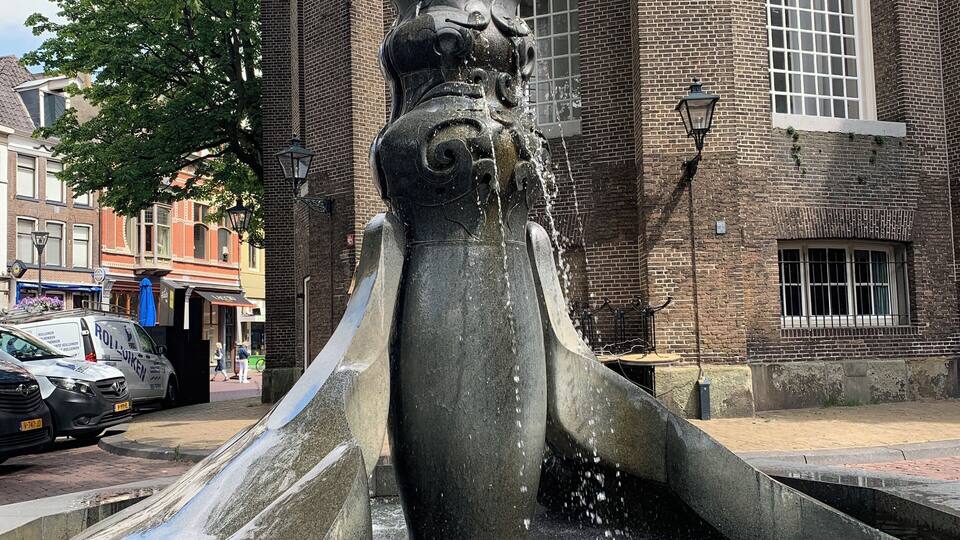 A stone fountain with water spouting from its center, set against a brick building with arched windows.