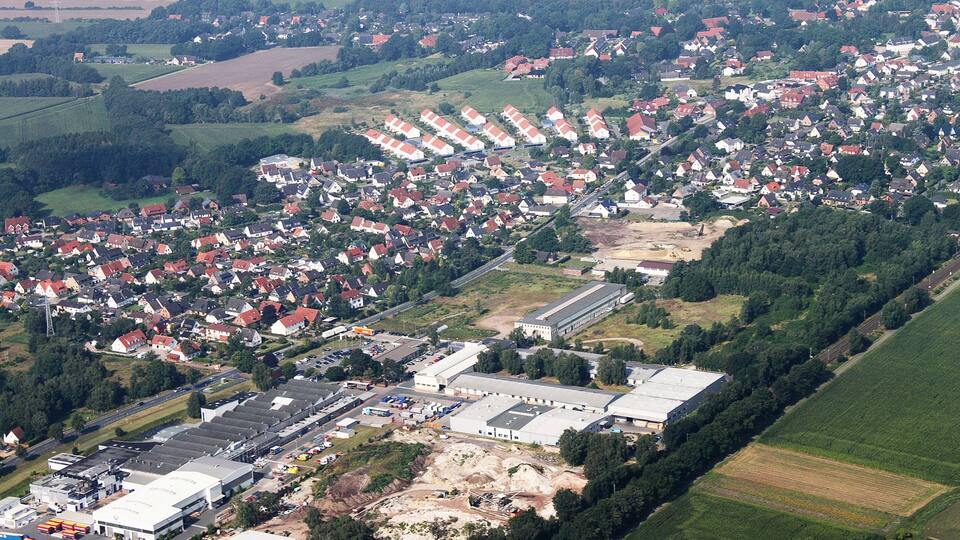 An aerial view of a residential area with houses, a large industrial building, and surrounding fields.