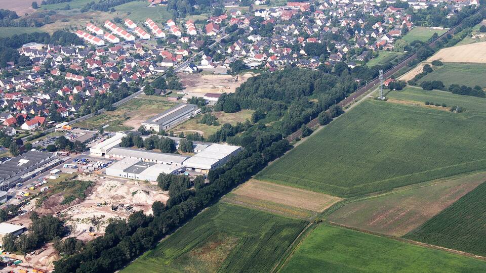 An aerial view of a residential area with houses, a large industrial building, and surrounding green fields.